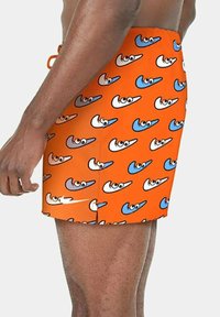 Uomo che indossa un costume da bagno arancione brillante con un motivo ripetuto di occhi dei cartoni animati e forme a giogo in blu, bianco e grigio.