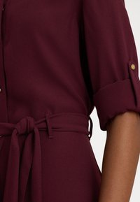 Bordové šaty s krátkymi vyhrnutými rukávmi, opaskom v páse a zlatými gombíkmi. Vyrobené z hladkej, ľahkej tkaniny s jemne textúrovaným povrchom.