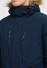 Tamsiai mėlynas parka su aukštu apykakle, kailinėmis kapišonu ir dviem užtrauktukais uždaromomis krūtinės kišenėmis. Yra reguliuojamos raišteliai ir lygus audinio paviršius.