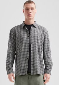 Zwart-witte geruite blouse met lange mouwen, een button-down kraag, knopen aan de voorkant en aansluitende mouwen. Gedragen over een zwart T-shirt.