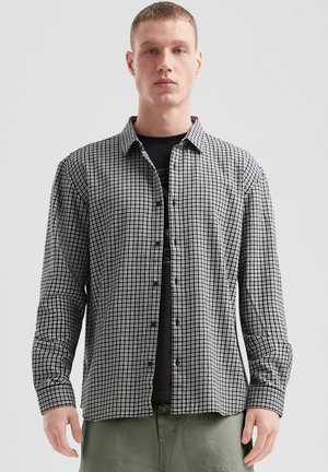 Zwart-witte geruite blouse met lange mouwen, een button-down kraag, knopen aan de voorkant en aansluitende mouwen. Gedragen over een zwart T-shirt.