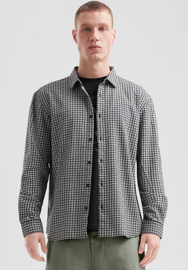 Zwart-witte geruite blouse met lange mouwen, een button-down kraag, knopen aan de voorkant en aansluitende mouwen. Gedragen over een zwart T-shirt.