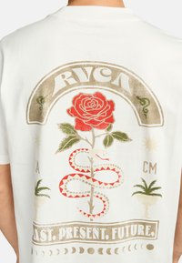 Camiseta de algodón blanca que presenta un gran diseño gráfico con una rosa roja, una serpiente verde, acentos dorados y el texto “PASADO. PRESENTE. FUTURO.”