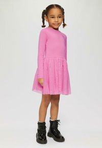 Rosa Langarmkleid mit einem Tüllrock; es hat ein tailliertes Oberteil, eine geraffte taille und schwarze Stiefel mit dicker Sohle und Schnürsenkeln.