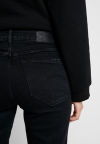 Svarta denimjeans med en figurnära silhuett, prydda med en läderlogotyp, bakfickor med sömmar och en subtilt urblekt yta.