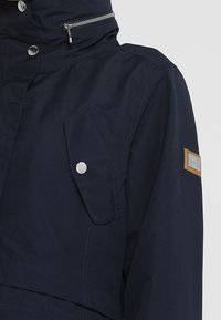 Veste bleu marine foncé avec col montant, fermeture éclair argentée et boutons-pression, poche à rabat sur la poitrine et un patch rectangulaire sur la manche supérieure.