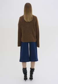 Brauner Strickpullover, weite blaue Jeans-Culottes und schwarze Absatzstiefel. Glattes Haar reicht über die Schultern. Minimalistischer Hintergrund.