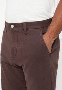 Vīrietis valkā tumši brūnus bikses ar priekšējo pogu un rāvējslēdzēju, baltu krekls iebāzts iekšā, un labā roka kabatā.