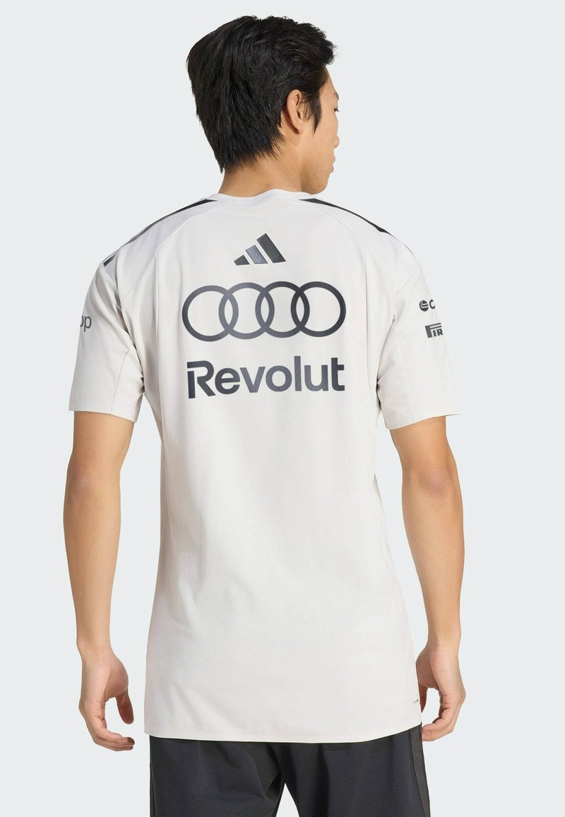 Man wearing white sports jersey with Adidas logo, Audi rings, and "Revolut" printed on the back, standing facing away against plain background.