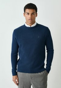 Marineblaues Strickpullover mit gerippter Textur und rundem Ausschnitt, getragen über einem weißen Hemd mit Kragen, kombiniert mit grauen Hosen.