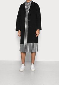 Manteau noir oversize avec un col à revers sur une robe chemise à rayures noir et blanc. Des baskets blanches complètent le look. Textures de tissu lisses.