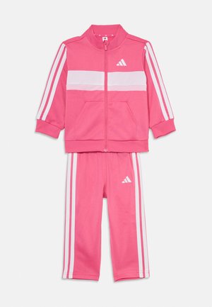 Chándal infantil rosa con franjas blancas en las mangas y pantalones, con chaqueta con cremallera y logo de Adidas en el pecho y el muslo.