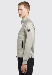 Lichtgrijze jas met een gevoerd ondergedeelte en een glad bovenste gedeelte. Voorzien van ritszakken en een logopatch op de linkermouw.