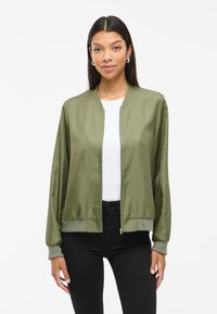 Olivovo zelená saténová bomber bunda so zipsom vpredu, rebrovanými manžetami a lemom; nosená cez biely top a čierne nohavice. Hladká textura.