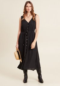 Zwarte maxi-jurk met een V-hals, mouwloos ontwerp, getailleerde taille en knoopdetails. Gestyled met een strohoed en zwarte kniehoge laarzen.