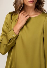 Olivgrüne Bluse mit einer weichen, glatten Textur, rundem Halsausschnitt und Puffärmeln. Verfügt über einen Knopfdetails am Ärmelaufschlag.