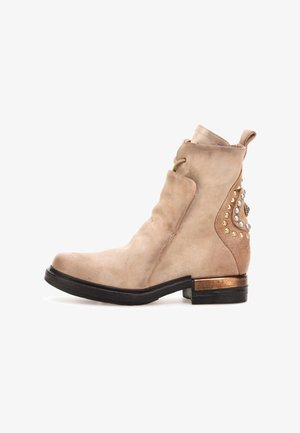 Stivale ankle in suede beige con punta arrotondata, borchie decorative sui lati e un accento metallico sul tallone. Texture liscia in generale.