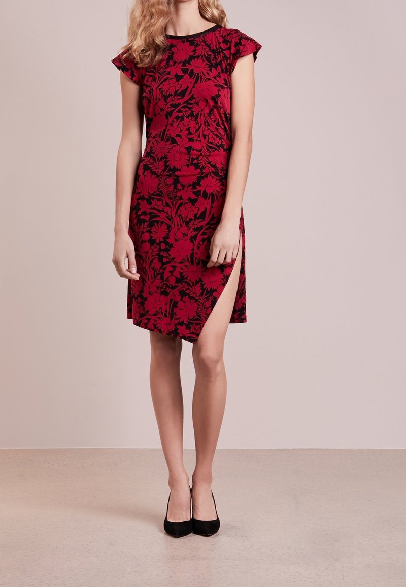 Femme portant une robe rouge longueur genou avec des motifs floraux noirs et une fente latérale haute jusqu'à la cuisse, associée à des chaussures à talons noirs.
