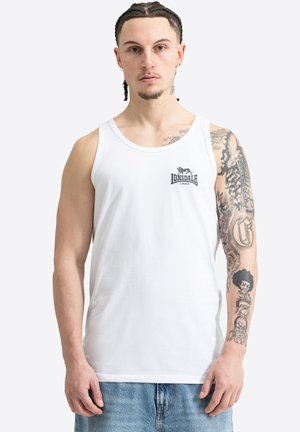 Uomo con capelli intrecciati e tatuaggi che indossa una canottiera bianca Lonsdale London e jeans blu, in piedi davanti a uno sfondo semplice.