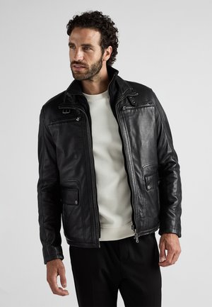 Mand med krøllet hår iført en sort læderjakke over en hvid sweater og sorte bukser, som kigger til siden mod en ensfarvet baggrund.