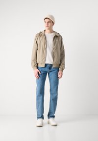Letvægts khaki vindjakke med elastiske manchetter, parret med en hvid T-shirt, blå jeans og hvide sneakers. Beige strikket beanie hue.