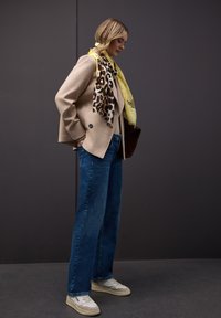 Femme portant une veste beige, un jean bleu, des baskets blanches et une écharpe jaune à motif léopard, debout devant un fond gris foncé.