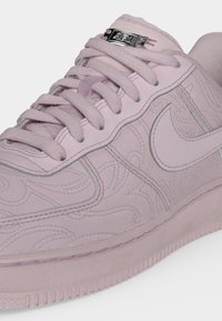 Ανοιχτό ροζ παπούτσι Nike με ανάγλυφα λουλουδάτα σχέδια, χοντρά κορδόνια και ασημένια ετικέτα κορδονιών στη γλώσσα σε απλό φόντο.