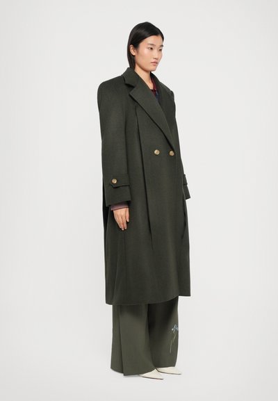 Manteau croisé vert avec de grands revers, boutons dorés et coupe légèrement surdimensionnée. Assorti à un pantalon à jambes larges et des chaussures blanches.