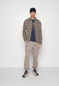 Chemise en flanelle à carreaux bleue et orange, t-shirt graphique marine, joggers beiges et baskets multicolores. Casquette noire sur la tête.