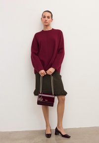 Bordový oversize sveter, olivovo zelená sukňa s třásňami a lesklá bordová kabelka so zlatými kovovými doplnkami. Masívne lesklé bordové topánky na hrubej podpätku.