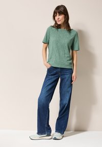 Chemise verte texturée à manches courtes avec un motif géométrique, associée à un jean bleu à jambes larges et des baskets de couleur claire sur un fond neutre.