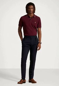 Άνδρας που φοράει μπορντώ πόλο, ναυτικές ριγέ παν trousers, καφέ δερμάτινα παπούτσια, στέκεται με το χέρι στην τσέπη πάνω σε απλή φόντο.