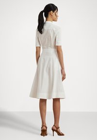 Biele šaty košeľového strihu s priliehavým živôtikom a rozšírenou sukňou, krátkymi rukávmi, gombíkovými detailmi a hladkou textúrou, doplnené hnedými sandálmi na podpätku.