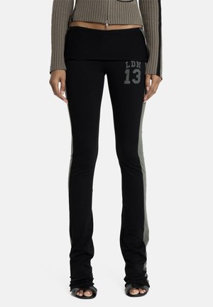 Nő fekete, bő szárú nadrágot visel szürke oldalsávval és "LDN 13" felirattal a bal combján, bézs színű, rövid, cipzáras pulóverrel párosítva.