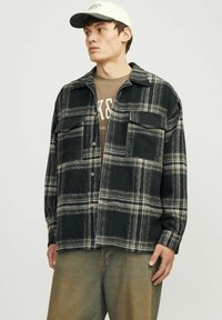 Sort-hvid ternet flannel skjorte med to brystlommer, knaplukning og krave, båret over en brun grafisk t-shirt og beige bukser.