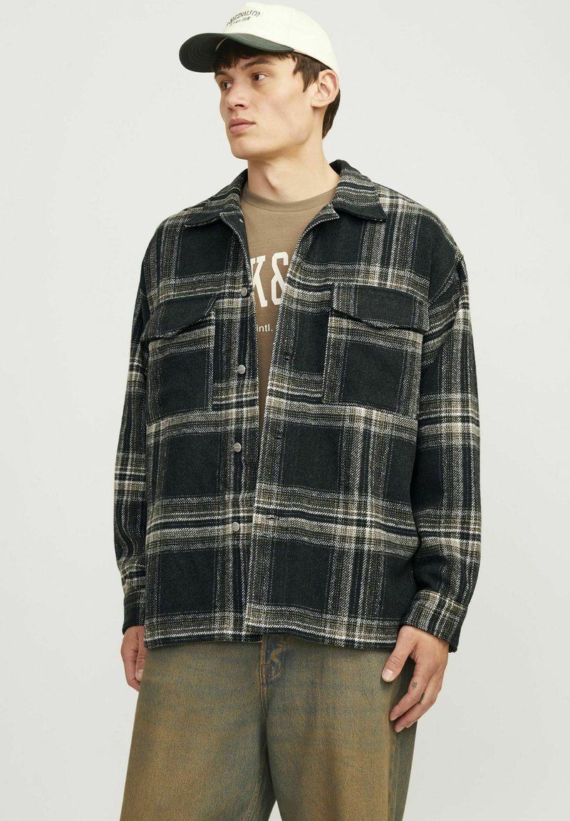 Sort-hvid ternet flannel skjorte med to brystlommer, knaplukning og krave, båret over en brun grafisk t-shirt og beige bukser.