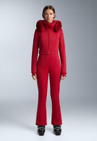 Piros síruházat cipzáros elülső r�ésszel, prémmel bélelt kapucnival, övvel a deréknál és bővülő szárakkal. Simább, rugalmas anyagból készült, varrási részletekkel.