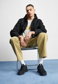 Um hoodie preto com fecho éclair sobre uma camisa branca, calças bege e sapatos de desporto pretos. O modelo está sentado numa cadeira cromada contra um fundo claro.