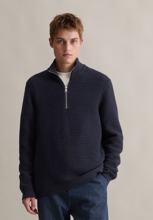 Navy Zip-Up-Pullover mit strukturiertem Waffelmuster und geripptem Saum, ausgestattet mit einem hohen Kragen und langen Ärmeln. Über einem weißen Hemd getragen.