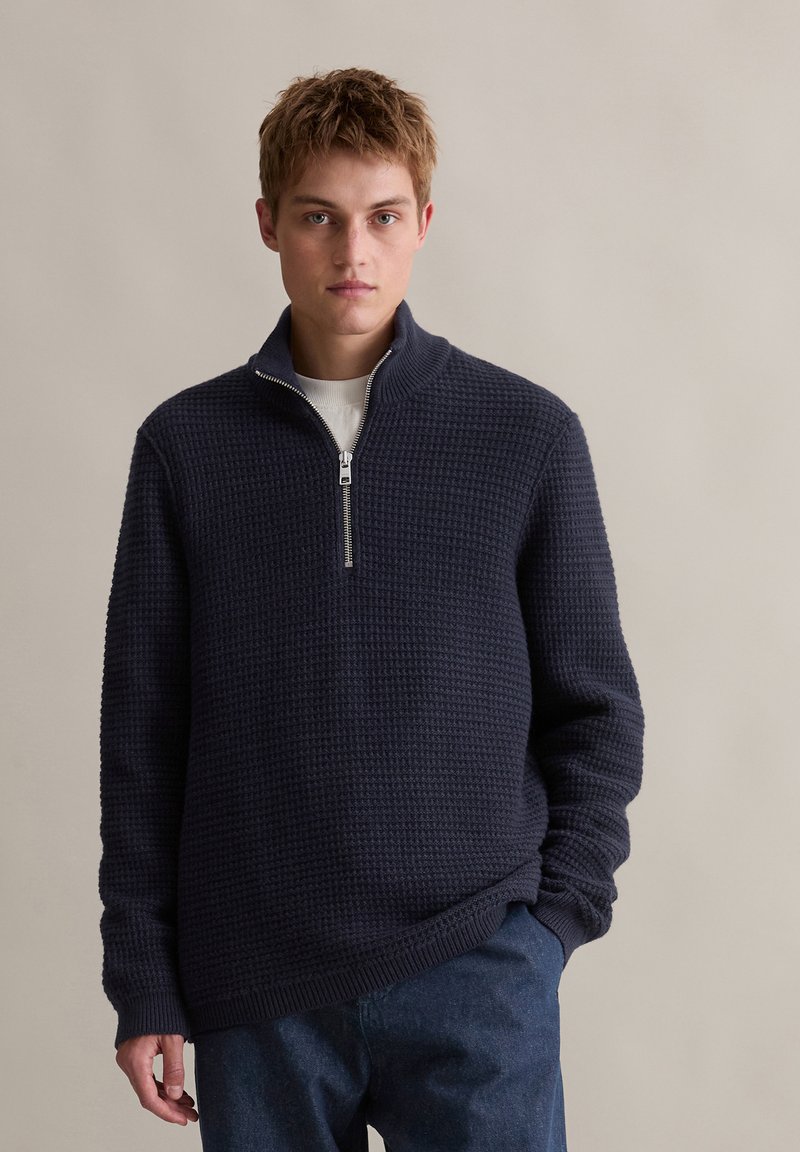 Felpa con zip di colore navy, con un motivo a waffle testurizzato e orlo a costine, dotata di colletto alto e maniche lunghe. Indossata sopra una camicia bianca.