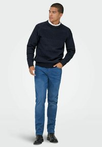Pull en tricot navy avec des motifs texturés et des poignets à côtes, associé à un jean bleu clair et des bottes noires. Le mannequin se tient avec une main dans la poche.
