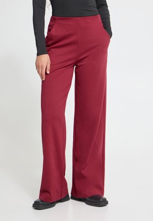 Persoon die hoge taille wijde bordeauxrode broek draagt met handen in de zakken en zwarte schoenen, gecombineerd met een aansluitende zwarte top met lange mouwen.