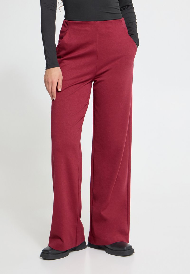 Personne portant un pantalon bordeaux taille haute à jambes larges, mains dans les poches, avec des chaussures noires, assorti à un haut noir cintré à manches longues.