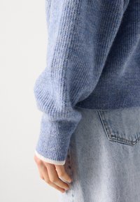 Blue knitted sweater with ribbed texture, featuring a white cuff accent. Paired with light denim jeans, showing a relaxed fit.