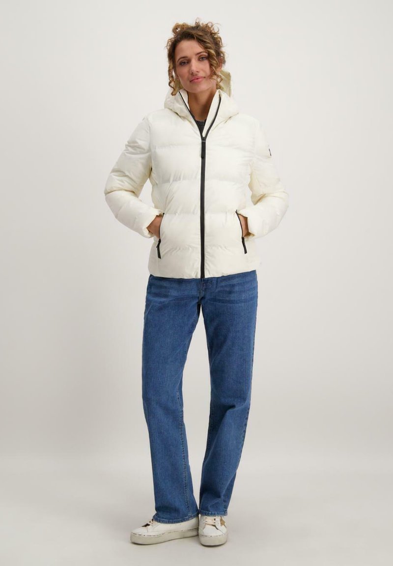 Witte pufferjack met capuchon, een voorritssluiting en twee zijzakken. Gecombineerd met blauwe jeans en sneakers, staande tegen een neutrale achtergrond.