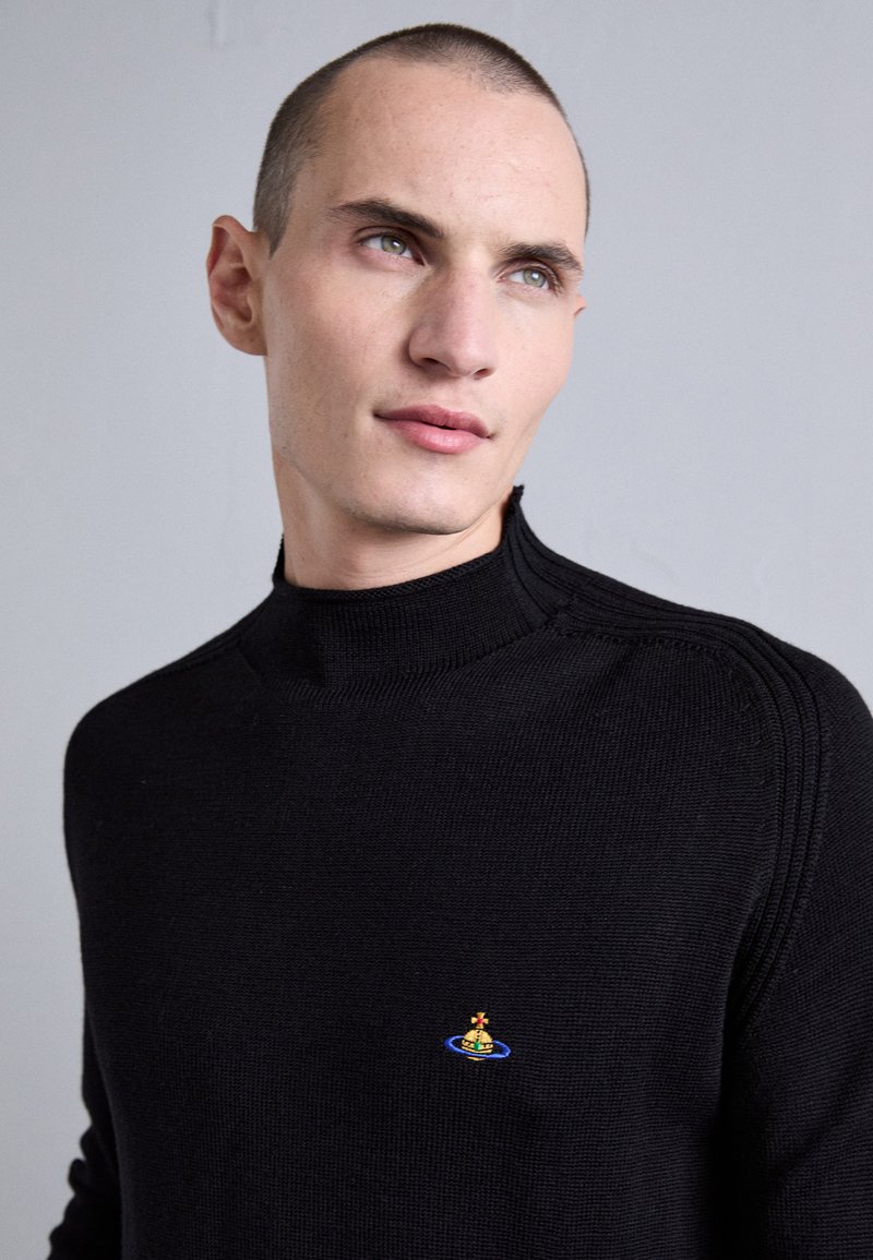 Junger Mann mit kurzen Haaren, der einen schwarzen Pullover mit hohem Kragen und einem kleinen bunten, orbähnlichen Emblem auf der Brust trägt, vor einem einfarbigen grauen Hintergrund.