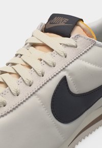 Μπεζ και λευκό αθλητικό παπούτσι Nike με κρέμ κορδόνια, μαύρο λογότυπο Nike στο πλάι και μαύρο σήμα λογότυπου στη γλώσσα.
