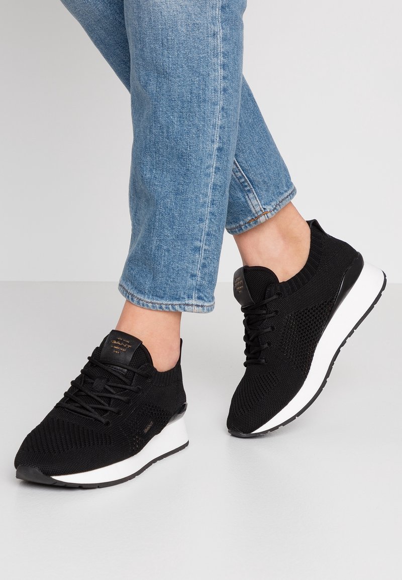 Black knitted sneakers featuring a lace-up design, white midsole, textured surface, and a rounded toe. Worn with light blue jeans.