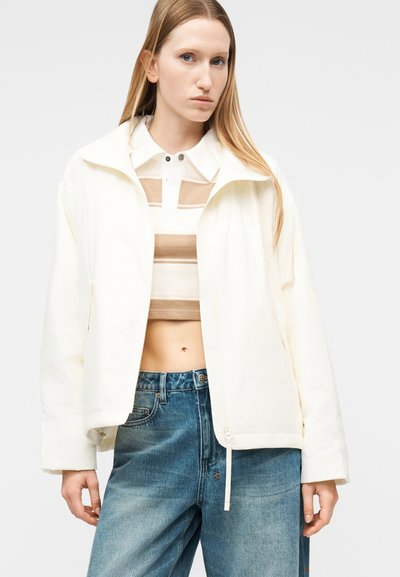 Γυναίκα με μαλλιά μακριά ξανθά, που φοράει λευκό μπουφάν πάνω από τοπ με μπεζ και λευκές ριγέ γραμμές, κομπόστο και ανοιχτόχρωμα μπλε τζιν, στέκεται μπροστά σε ένα απλό φόντο.