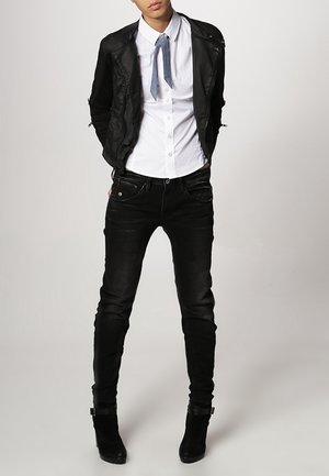 Persona con camisa blanca, chaqueta corta negra, vaqueros ajustados negros, botines negros y corbata azul sobre un fondo liso.